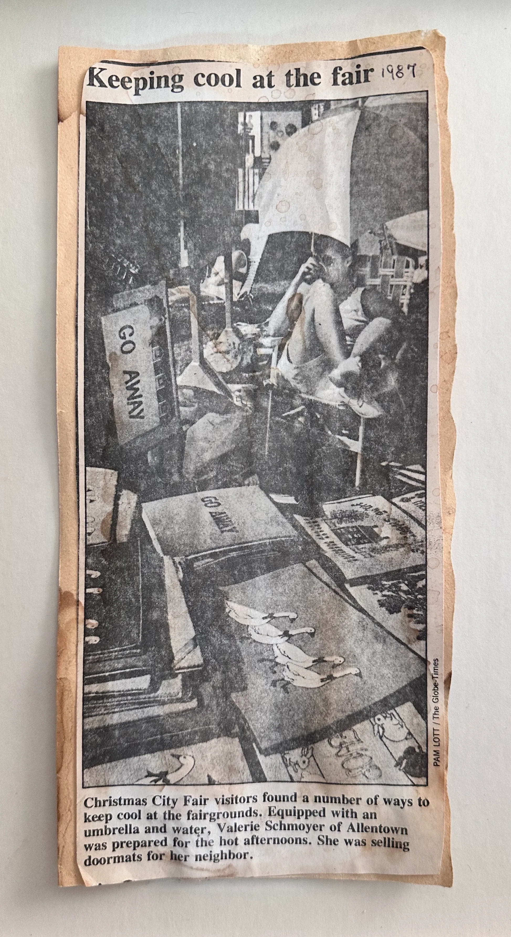 1987 Bethlehem Globe-Times newspaper clipping. Headline: Keeping cool at the fair. Photo: Val on folding chair at booth with novelty doormats sipping frim a cup, sunglasses on, legs folded in. Caption: Christmas City Fair visitors found a number of ways to keep cool at the fairgrounds. Equipped with an umbrella and water, Valerie Schmoyer of Allentown was prepared for the hot afternoons. She was selling doormats for her neighbor.